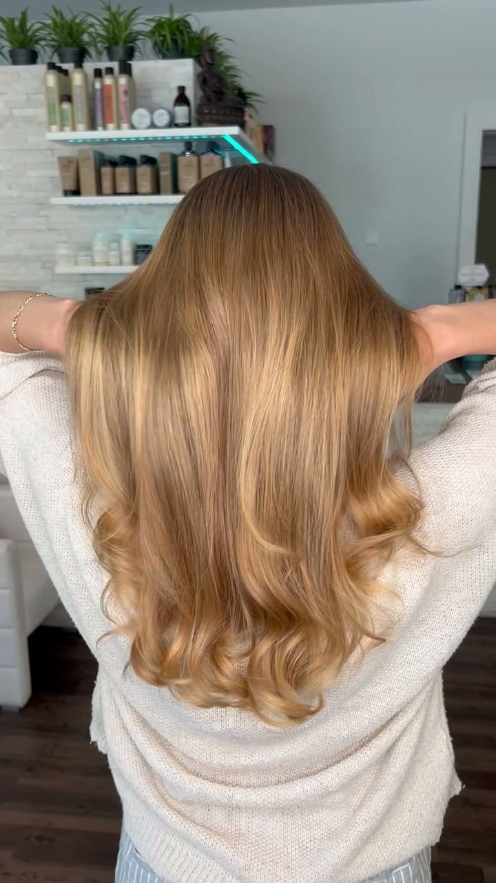 Rückansicht einer Kundin mit langem, honigblondem Haar in weichen Wellen: Sie hebt das Haar an, sodass Farbverlauf und Glanz in Bewegung sichtbar werden. Aufgenommen im Salon von Valesca Hair vor der Produktwand.